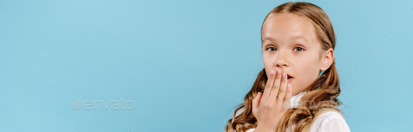 panoramic shot of shocked and cute kid looking at camera and obscuring face isolated on blue