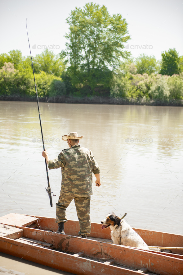 Slim male angler in uniform outfit standing in shabby boat with rod and dog,  fishing in morning. Stock Photo by halynabobyk