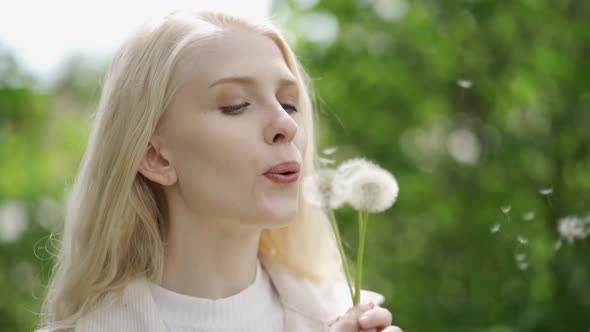 A Romantic Woman with Light Curls Fervently Blowing Dandelions