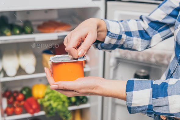 cropped view of woman opening yogurt near open fridge with fresh food on shelves