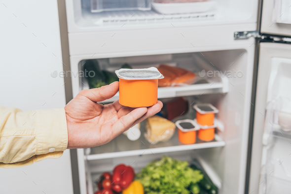 cropped view of man holding yogurt near open fridge full of food