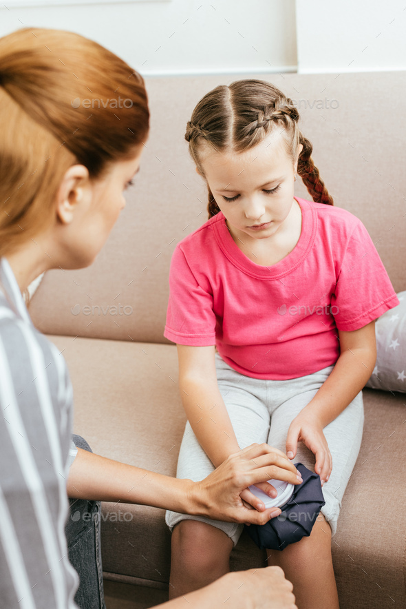 mom holding ice bag compress on painful knee of sad daughter