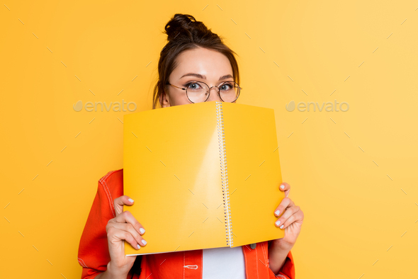 young student in glasses obscuring face with copy book while looking at camera isolated on yellow