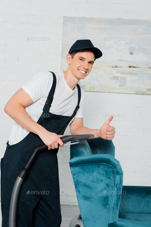 happy cleaner in cap showing thumb up while dry cleaning blue armchair