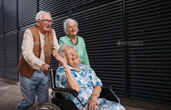 Elderly friends pushing senior woman in wheelchair.
