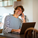 Smiling young man sitting at home talking on cell phone while working on laptop - PhotoDune Item for Sale