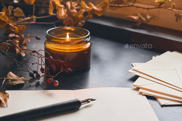 Cozy autumn still life fall leaves, sheets of paper, fountain pen and burning candle on windowsill.