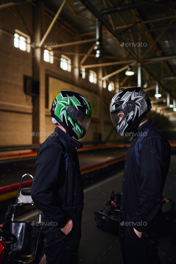 two go kart competitors in sportswear and helmets standing face to face on circuit, indoor karting