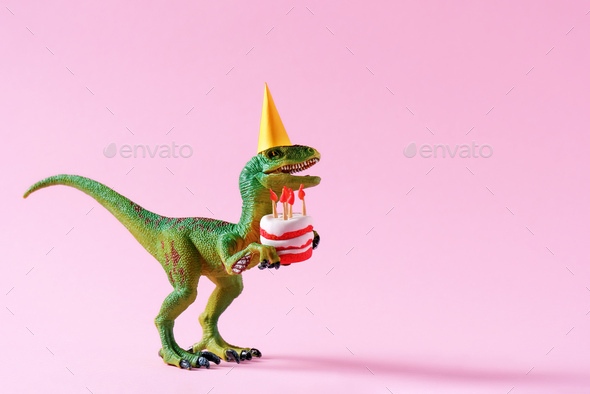 Cute happy green dinosaur in birthday hat holding cake with flaming candles on pink background.