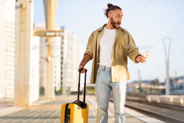 Shocked sad millennial caucasian man with suitcase looks on phone at train station