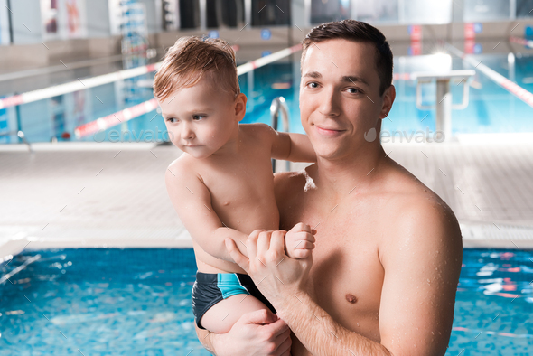 happy swim trainer holding in arms toddler kid near swimming pool