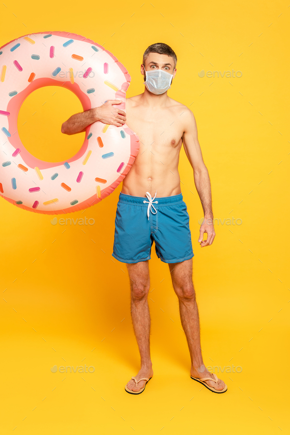 full length view of guy in swim trunks and medical mask with swim ring on yellow