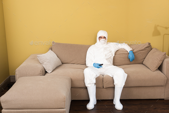 Man in hazmat suit and medical mask holding remote controller on couch
