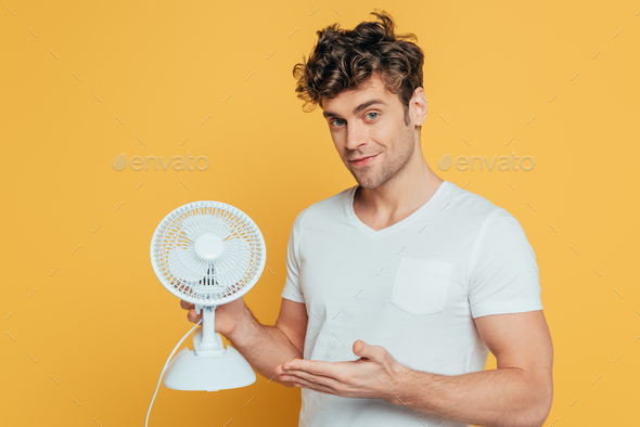 Man smiling, looking at camera and pointing at desk fan isolated on yellow