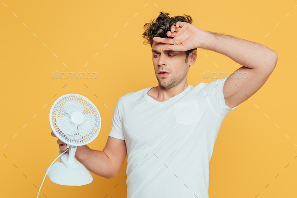 Confused man with raised hand looking at desk fan isolated on yellow