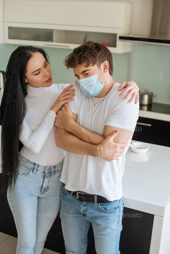 beautiful girl hugging cold ill boyfriend in medical mask at home during self isolation