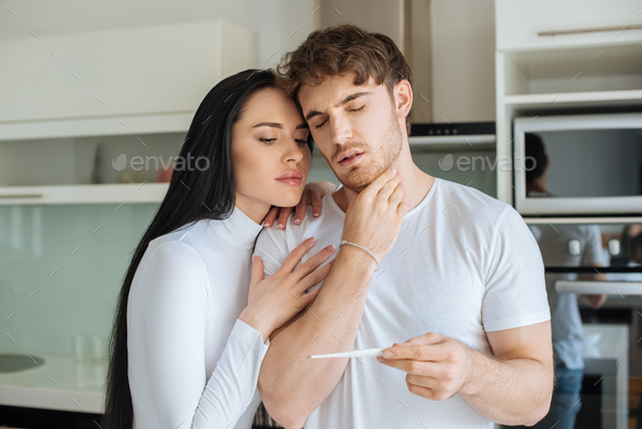 Worried girl hugging boyfriend with sore throat and looking at thermometer during self isolation