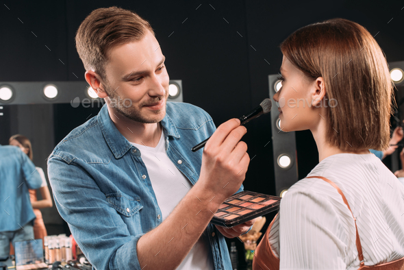 Smiling makeup artist applying blush on young model in photo studio