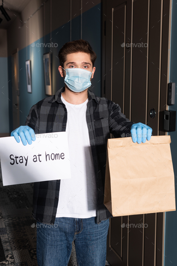 Courier in medical mask and latex gloves holding card with stay at home lettering and package on