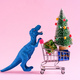 Happy blue dinosaur carrying shopping cart with Christmas tree and gift boxes on pink background. - PhotoDune Item for Sale