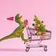 Happy green toy dinosaur carrying shopping cart with Christmas tree on pastel pink background.  - PhotoDune Item for Sale