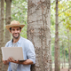 A young man wearing a straw hat leans against a tree with a laptop in his hand. - PhotoDune Item for Sale
