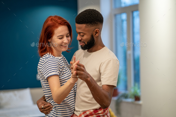 Loving young diverse couple dancing romantic dance in modern bedroom with panoramic windows.