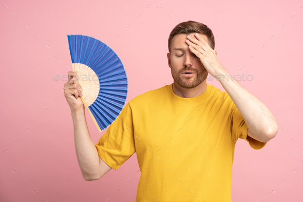 Sweaty tired man touching forehead using paper fan suffer from heat, cooling in hot summer weather