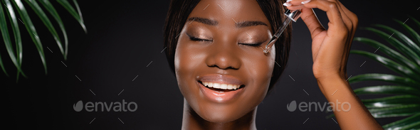 African American Woman Applying Serum On Face Near Green Palm Leaves Isolated On Black