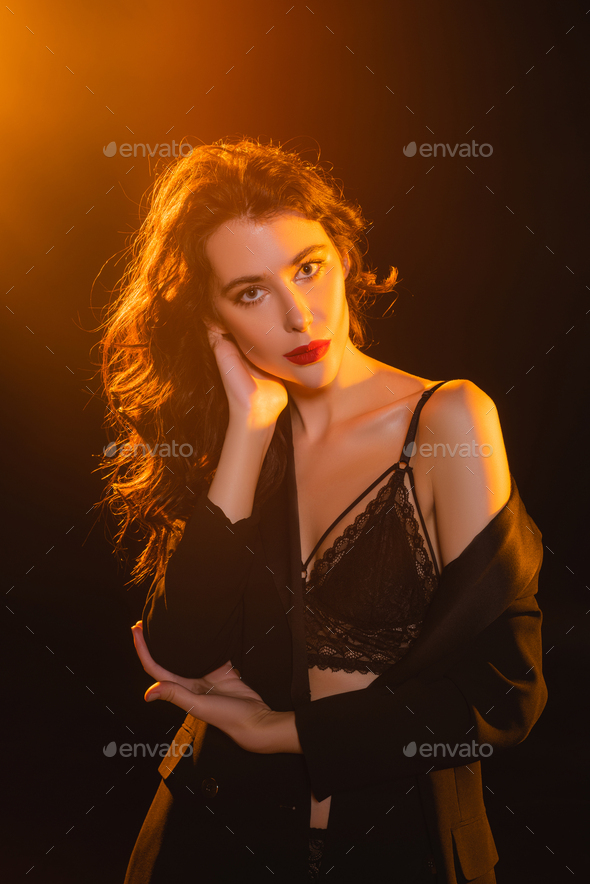 seductive and curly woman in lace bra and blazer looking at camera on black  Stock Photo by LightFieldStudios