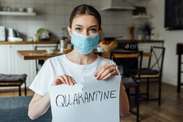 Girl in medical mask tearing card with quarantine lettering at home