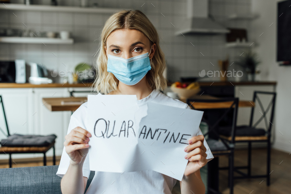 Woman in medical mask tearing card with quarantine lettering at home