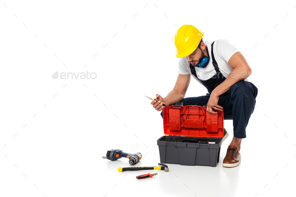 Workman in hardhat and ear defenders holding screwdriver near tools and toolbox on white background