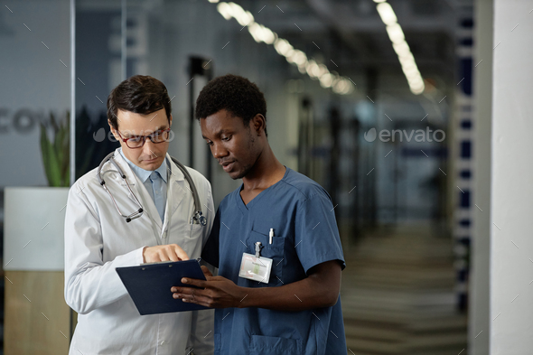Confident mature physician in lab coat pointing at medical document in clipboard