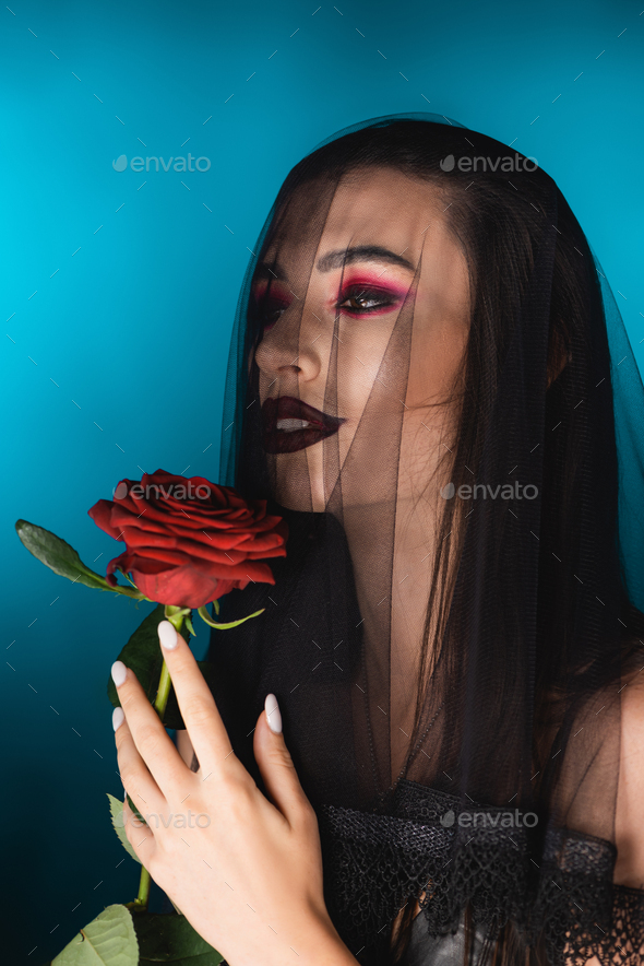 evil bride with black makeup looking away through veil and holding rose on blue