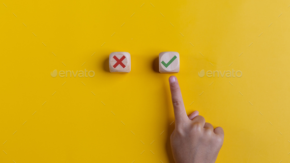 Right and wrong icons on wooden cubes with hand choosing the right icon on yellow background.
