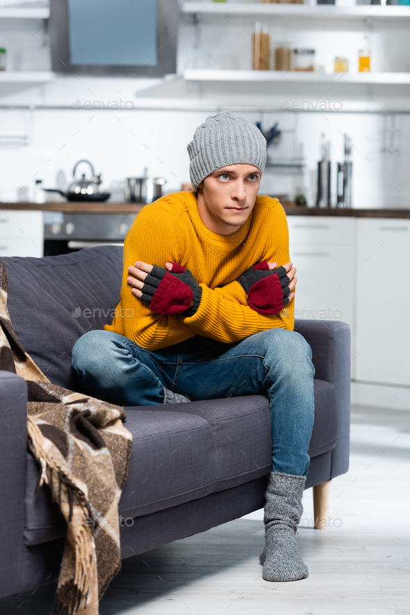 freezing man in warm hat and fingerless gloves hugging himself while sitting on sofa in cold kitchen
