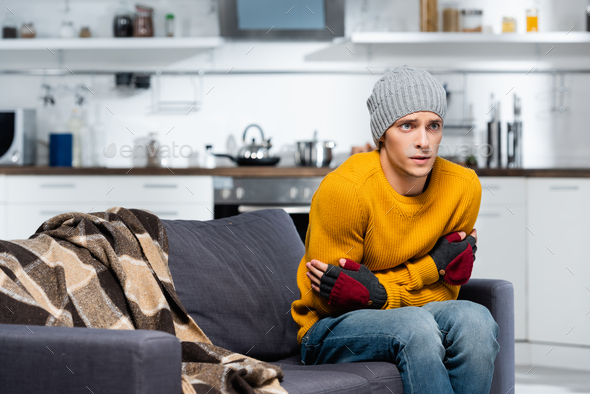 young man in warm hat and fingerless gloves hugging himself while sitting on sofa in cold kitchen