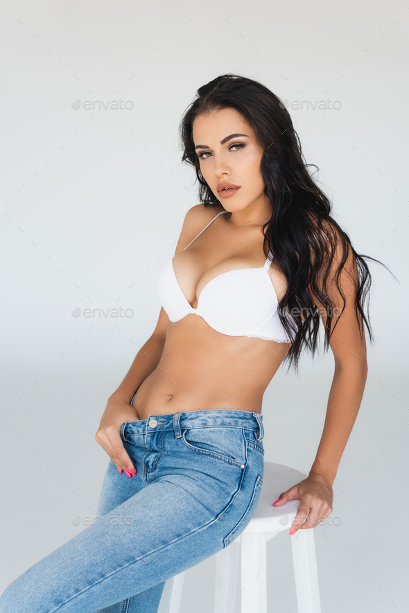 brunette woman in jeans and bra sitting on stool on grey Stock Photo by  LightFieldStudios
