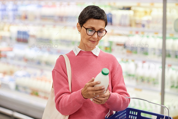 Mature woman holding bottle of milk in supermarket checking expiration date