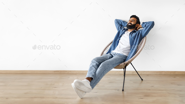 Indian man relaxing with hands behind head in armchair indoor - Stock Photo - Images