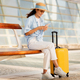 Glad young caucasian lady in hat with suitcase typing on smartphone, waiting for transport - PhotoDune Item for Sale