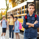 Upset schoolboy standing alone near school bus while classmates chatting on background - PhotoDune Item for Sale