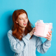 Curious teen girl with red hair, shaking gift box and wonder what inside, standing over blue - PhotoDune Item for Sale