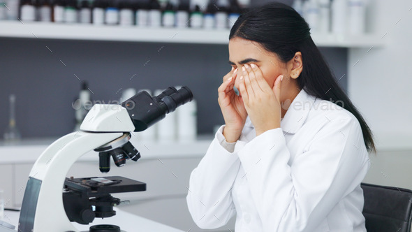 Female scientist looking through microscope and rubbing her eyes while suffering from eye strain or