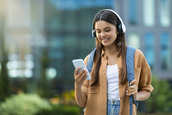 Cute woman college student using headphones and smartphone outdoors