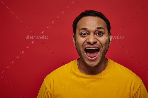 positive emotion, excited indian man in yellow t-shirt with opened mouth on red background