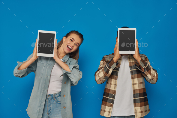 african american man obscuring face with digital tablet near excited woman on blue backdrop