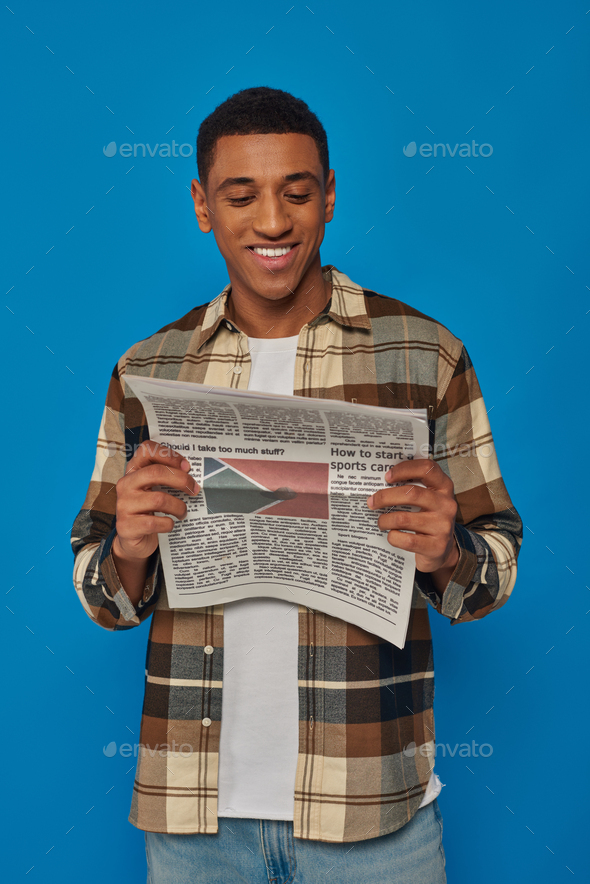 cheerful african american man in plaid shirt reading printed newspaper and smiling on blue backdrop