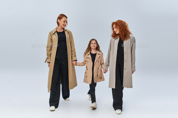 three generations, redhead women and girl in trench coats holding hands and walking on grey backdrop - Stock Photo - Images